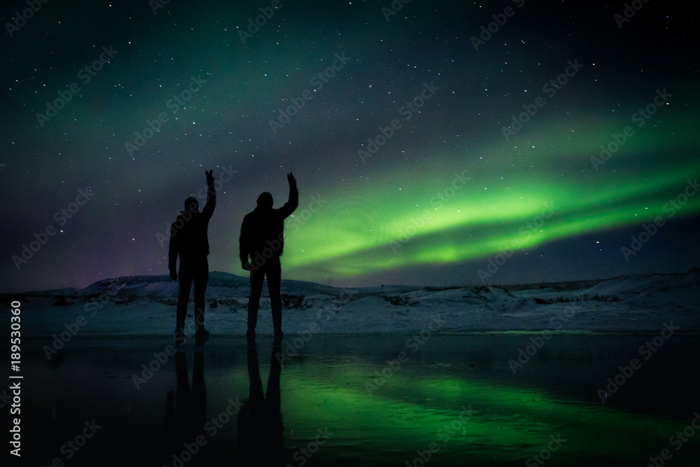 two men silhouettes raise their hands in a peace sign in front of the Northern Lights in Iceland