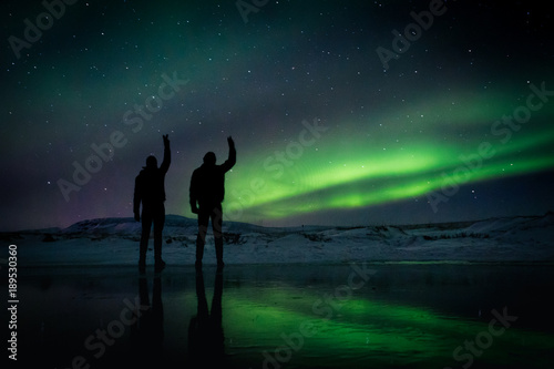 two men silhouettes raise their hands in a peace sign in front of the Northern Lights in Iceland