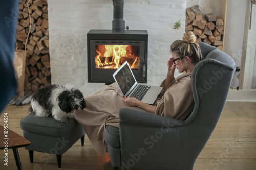 Beautiful middle-aged woman next to the fireplace relaxes in the living room and works on laptop from house. Girl in 30's surveyed for the  home bussiness project