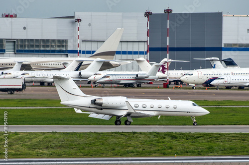 White private business jet taxiing on runway after landing. A lot of difference business jets on parking places on background.