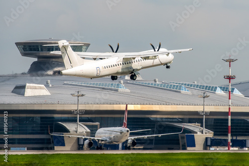 Regional turboprop twin-engine white passenger airplane taking off. Airport terminal at background.