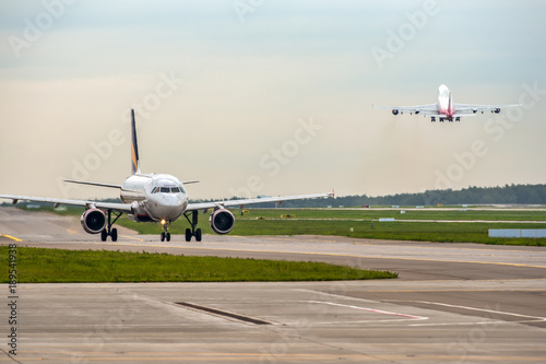 Modern passenger airplane taxiing while big widebody aircraft taking off at airport.