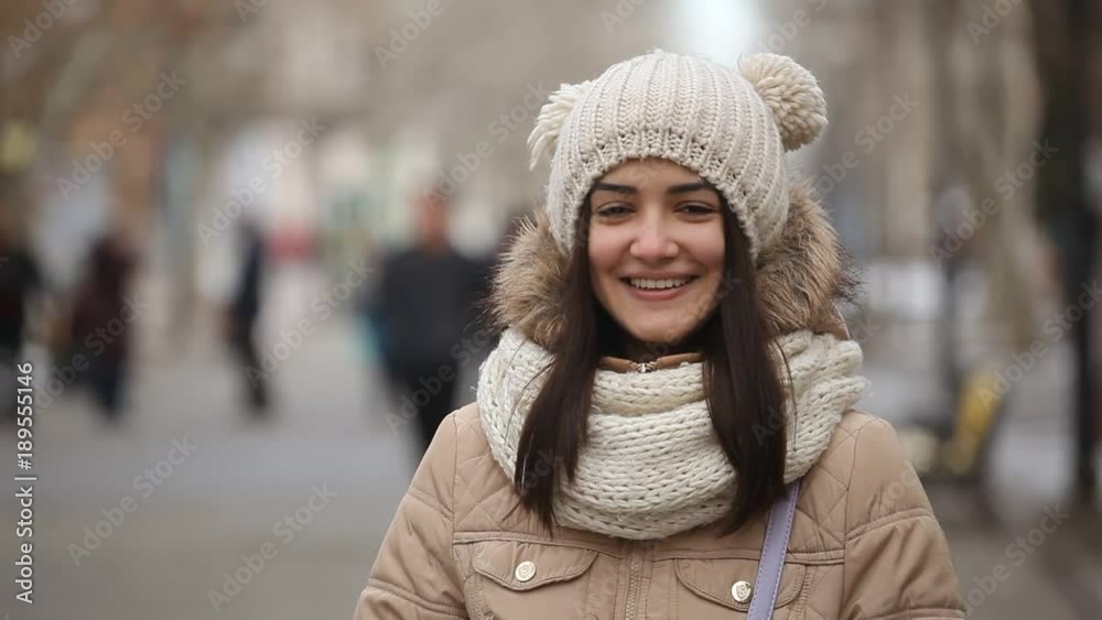 An optimistic view of a smiling girl with long hair in an anorak and a knitted hat with funny balls who stands on an alley in winter An optimistic view of a smiling girl with long hair in an anorak and a knitted hat with funny balls who stands on an alley in winter