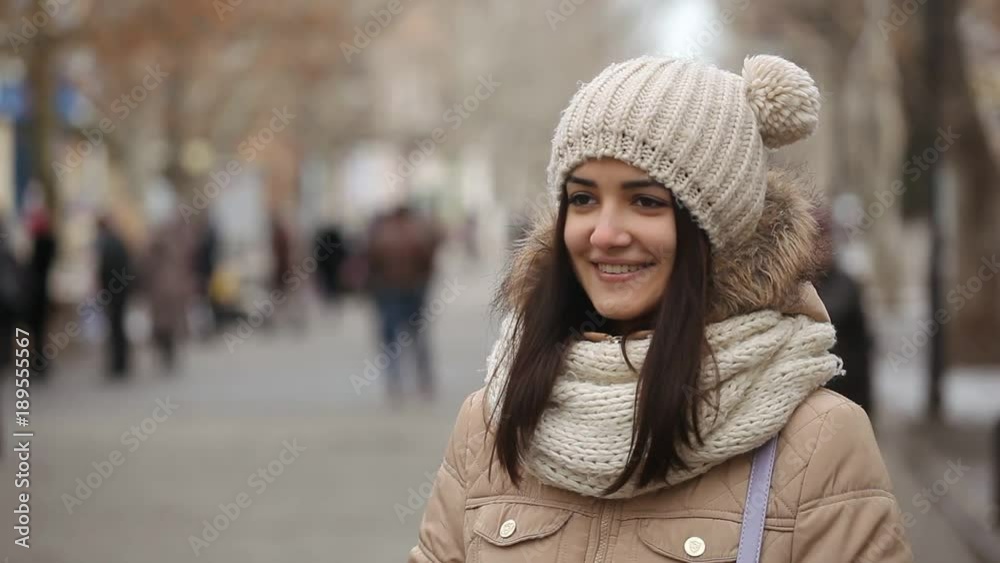 A cheery view of a smiling girl with long hair in an anorak and a knitted hat with two funny balls who stands on an alley in winter