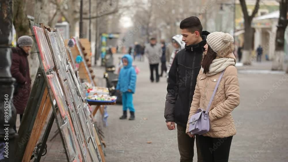 An amazing view of a lovely young couple looking at stands with pictures on a park alley in winter. A happy girl points at one painting. An amazing view of a lovely young couple looking at stands with pictures on a park alley in winter. A happy girl points at one painting.