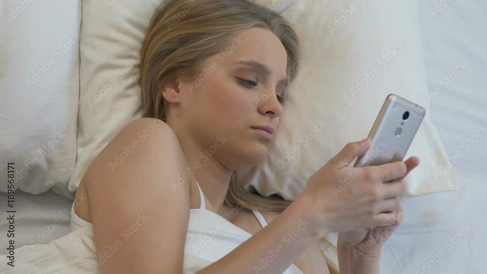 Sleepy young woman setting alarm in telephone and putting it under the pillow Sleepy young woman setting alarm in telephone and putting it under the pillow