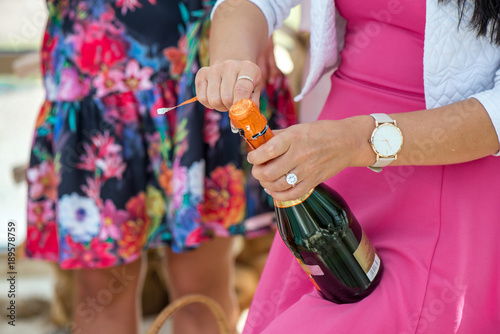 Girl in pink dress is opening champagne bottle to pour the drink in glasses. Cork is about to pop out. Cheerful bride and bridesmaids party 