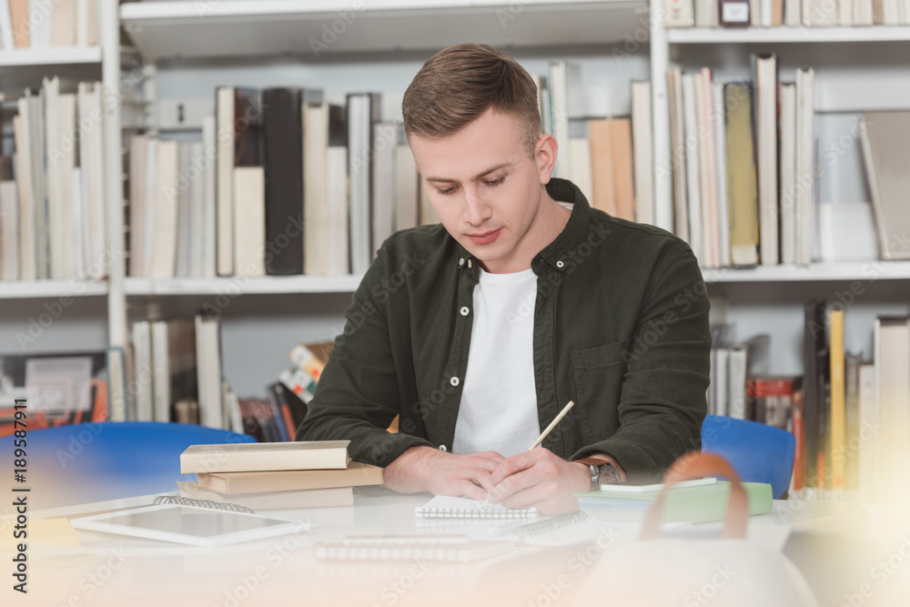 male student writing something to notebook in library male student writing something to notebook in library