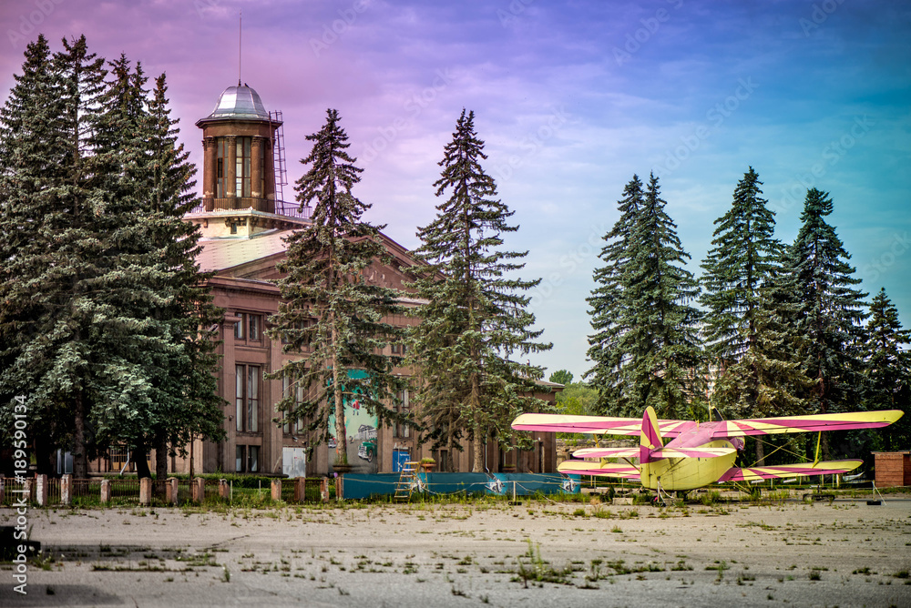 Obraz premium An old yellow and pink plane in an abandoned airport next to dark forest in an overcast day. Military plane, old russian aircraft. Soviet mass-produced single-engine biplane at an field aerodrome