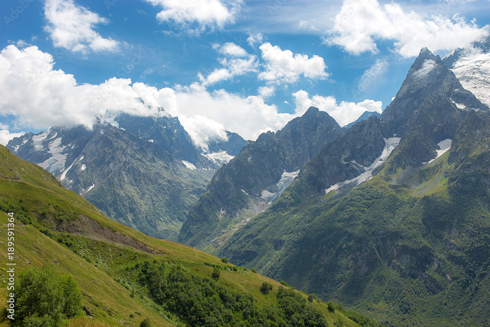 Obraz premium summer landscape of the Caucasus mountain peaks, green slopes in the grass with snow-capped peaks against the blue sky with clouds