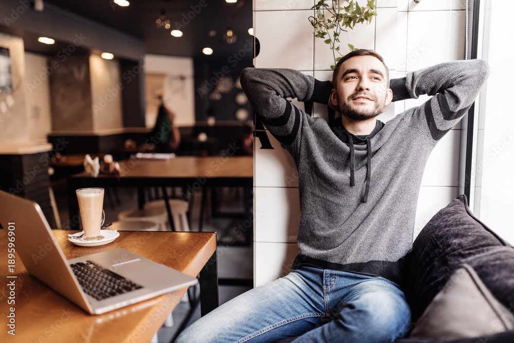 Fototapeta premium Relaxed young man holding hands behind head and smiling while sitting at the table with laptop in modern cafe