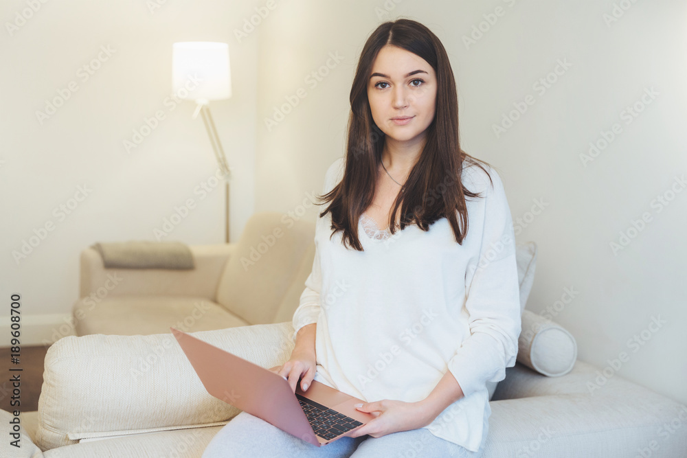Pretty young brunette female relaxes at sofa in spacious white room, watches short video on portable laptop computer, connected to wireless internet, spends free time at home. People and lifestyle