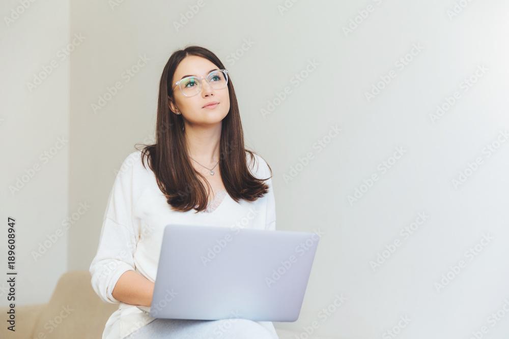 Naklejka premium Pensive young brunette female sits in front of opened laptop, thinks about interesting presentation, wants to impress everyone by her work, wears spectacles, sits against white wall