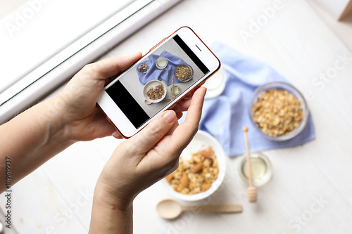 Close up of female hands holding mobile phone taking a picture of granola cereals, milk yogurt and trail mix vegetarian dieting breakfast for a new food blog article. Top view, overhead, copy space.