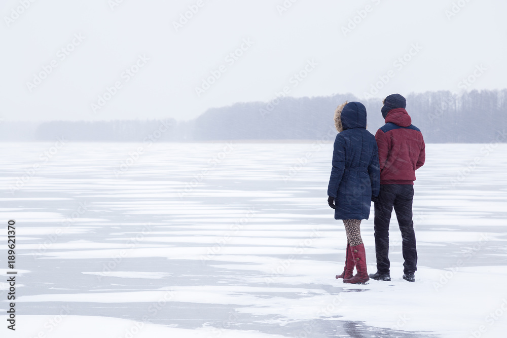 Young couple in snow blizzard walking on the lake ice and staring forward. Cold atmosphere in the afternoon. Foggy air. Winter concept.