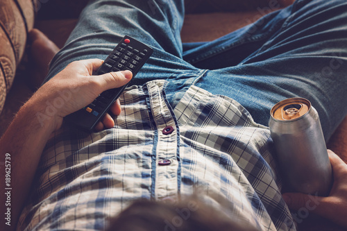 After work a guy lying on sofa, drinking a cold beer and watching sport tv channel. Hand holding a aluminum can and remote control. Man's resting time at home concept. 