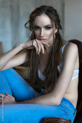 A beautiful thin girl wearing a white livery and unbuttoned jeans sits on an armchair in a loft-style room © Алексей Торбеев