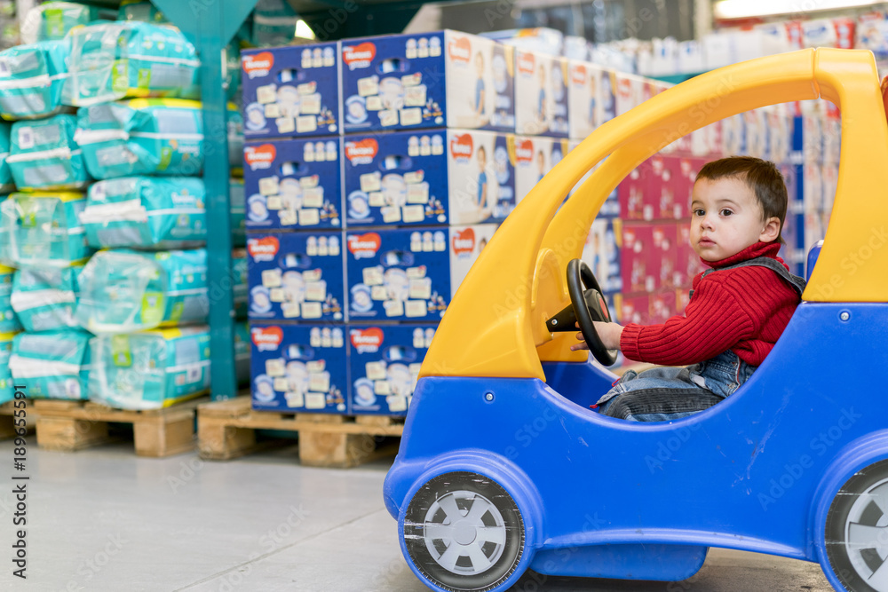 a little cheerful boy in red sweater in a children's car in a shopping center on a background of diapers a little cheerful boy in red sweater in a children's car in a shopping center on a background of diapers