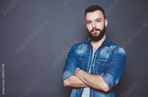 Handsome thoughtful Young bearded man in a denim shirt with arms crossed against a gray background.