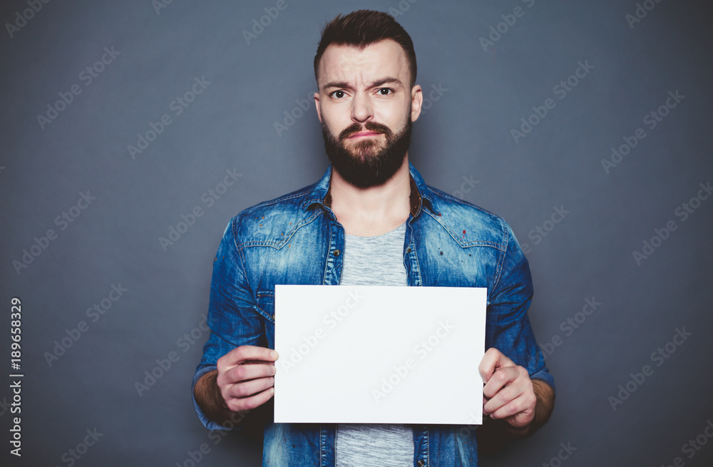 Everything is for you. Handsome young smiling man in a denim shirt shows a white sheet of paper in the camera on a gray background. Area for advertising. Everything is for you. Handsome young smiling man in a denim shirt shows a white sheet of paper in the camera on a gray background. Area for advertising.