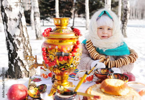 child girl in a fur coat and in a scarf in Russian style holding a large samovar in the hands of pancakes with red caviar, matryoshkas and utensils in Khokhloma style. Maslenitsa, Russia, winter