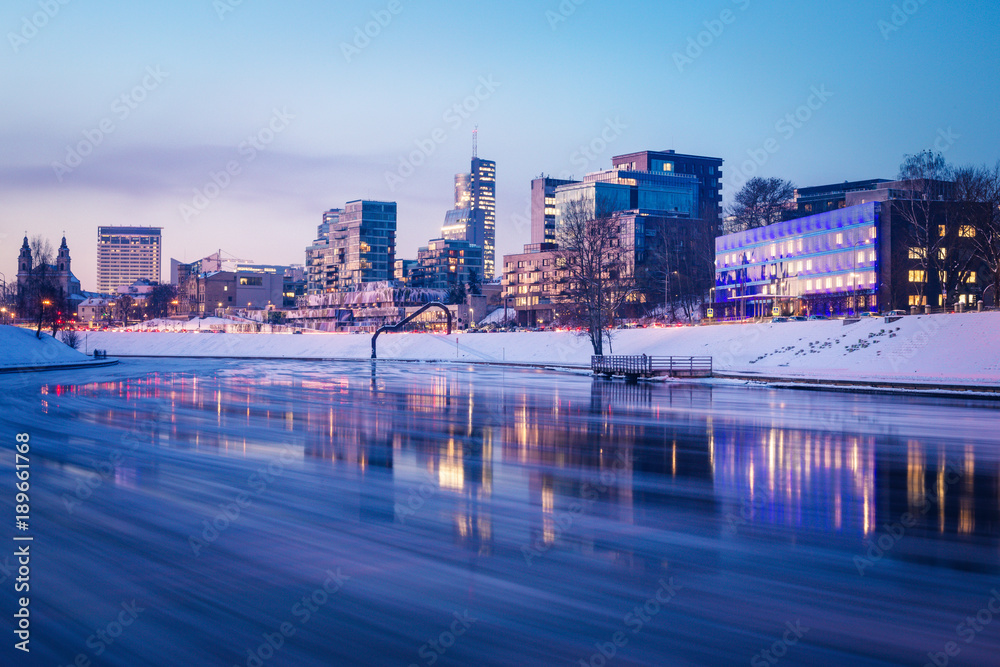 Fototapeta premium Cityscape skyline with office buildings on a river bank, clear blue sky, and reflections on the water during twilight blue hour in Vilnius, Lithuania