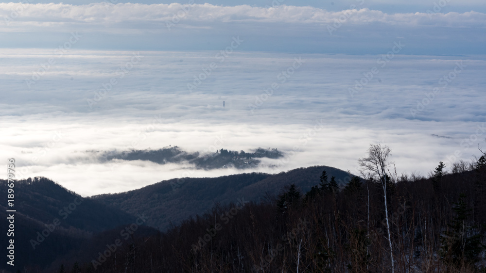 Landscape view from Sljeme or Medvednica mountain down on clouds covering the city of Zagreb in Croatia with a visible smoking heat plant chimney  above the clouds during sunrise