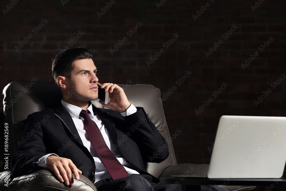 Handsome young man in suit talking by phone while sitting in armchair indoors Handsome young man in suit talking by phone while sitting in armchair indoors