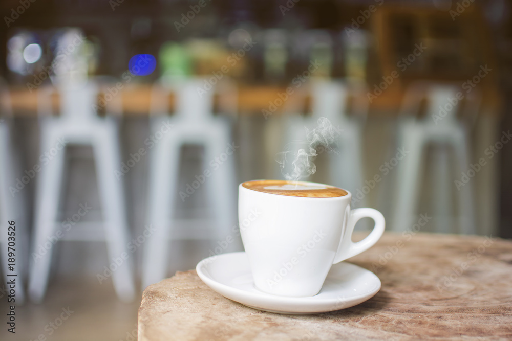White cup of hot steaming coffee on an old log in the cafe with blurred coffee bar background
