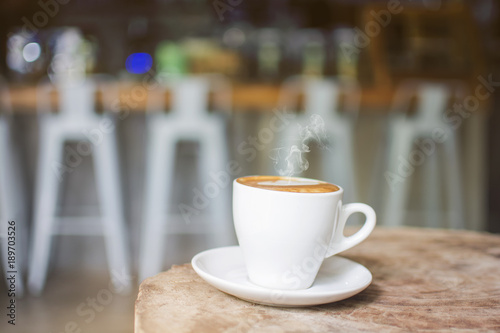 White cup of hot steaming coffee on an old log in the cafe with blurred coffee bar background