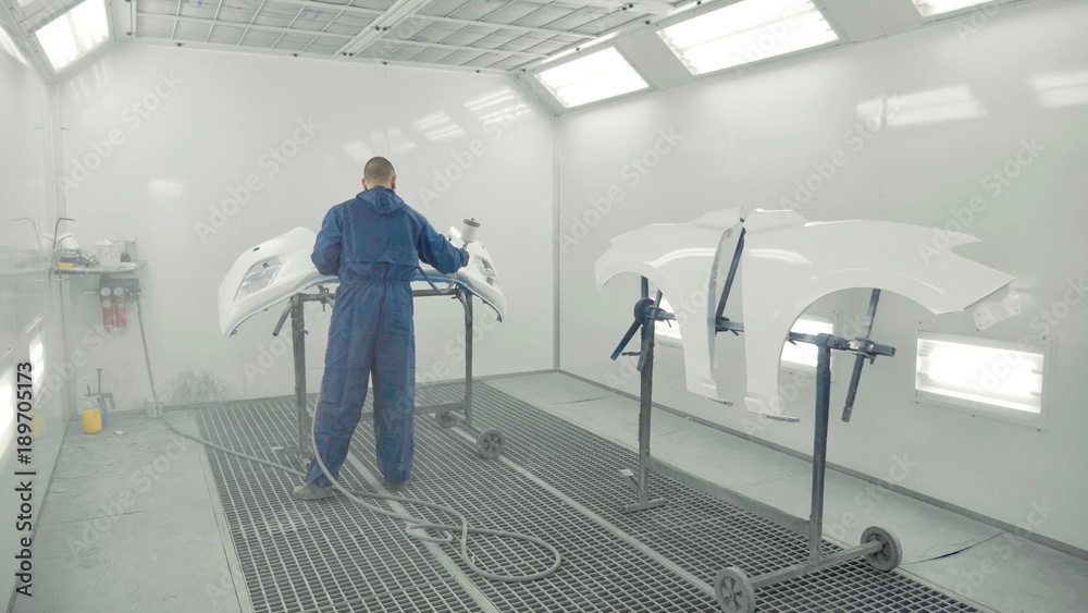 Auto painter spraying white paint on car front bumper in special booth