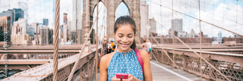 Happy woman using phone walking in NYC on Brooklyn Bridge. New York city lifestyle young Asian girl commuting or summer travel tourist on USA vacation. Banner panorama.