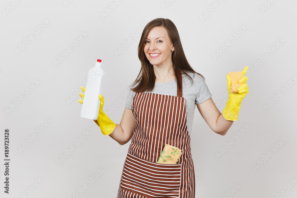 Young housewife in yellow gloves, striped apron, cleaning rag in pocket isolated on white background. Woman spreading hands, holding bottle with cleaner liquid, sponge. Copy space for advertisement.