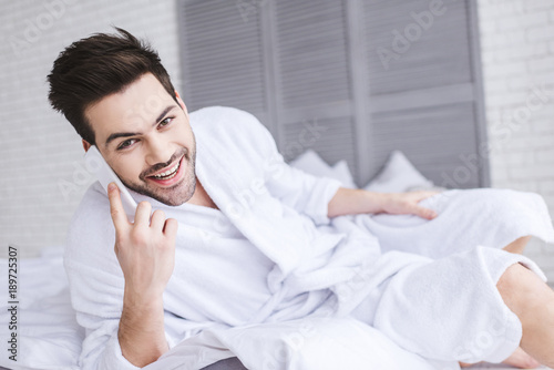 handsome young man in bathrobe talking by smartphone and smiling at camera