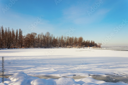 a frozen river by the shore of a forest under a blue sky © Nikolay