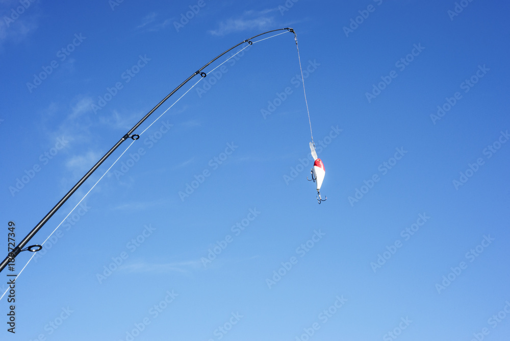 The spinning and brightly-colored crankbait are against the blue cloudless sky. The fishing equipment is on a cyan background. It is cast a line.