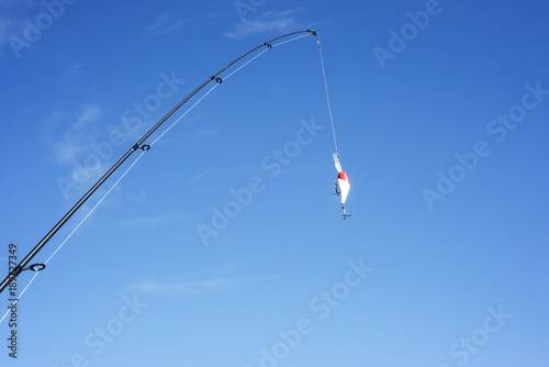 The spinning and brightly-colored crankbait are against the blue cloudless sky. The fishing equipment is on a cyan background. It is cast a line.