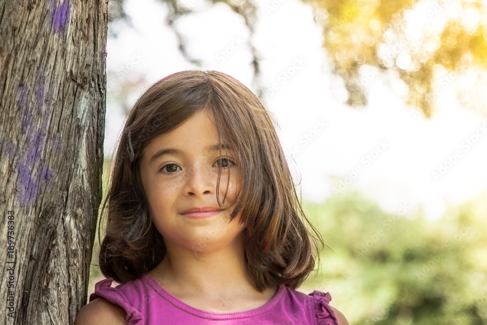 Happy young girl with pink t-shirt leaning back against a tree smiling and looking at camera in a park on a sunny summer day. Golden lens flare. Happy young girl with pink t-shirt leaning back against a tree smiling and looking at camera in a park on a sunny summer day. Golden lens flare.