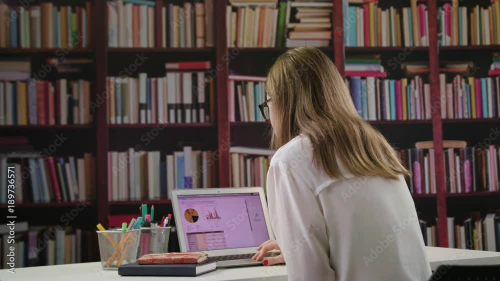 A young lady using a laptop and drinking coffee in the library. Medium. Dolly shot. A young lady using a laptop and drinking coffee in the library. Medium. Dolly shot.