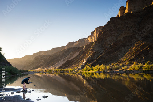 A hiker collecting water from a river reflecting the golden light of sunrise in the Fish River Canyon with high mountains reflected in  the calm water
