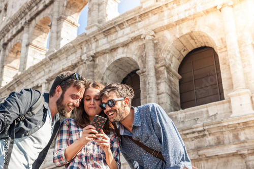 Photography Three happy young friends tourists at Colosseum in Rome using smartphone having fun on sunny day