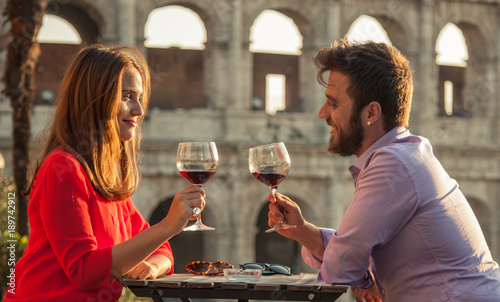 Photography Romantic couple drinking glass of red wine sitting at restaurant table in front of colosseum in rome at sunset