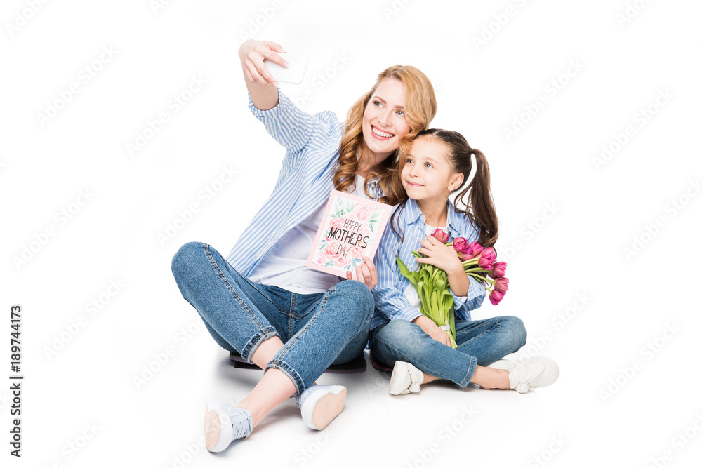 smiling mother with postcard and daughter with flowers taking selfie together isolated on white, mothers day holiday concept