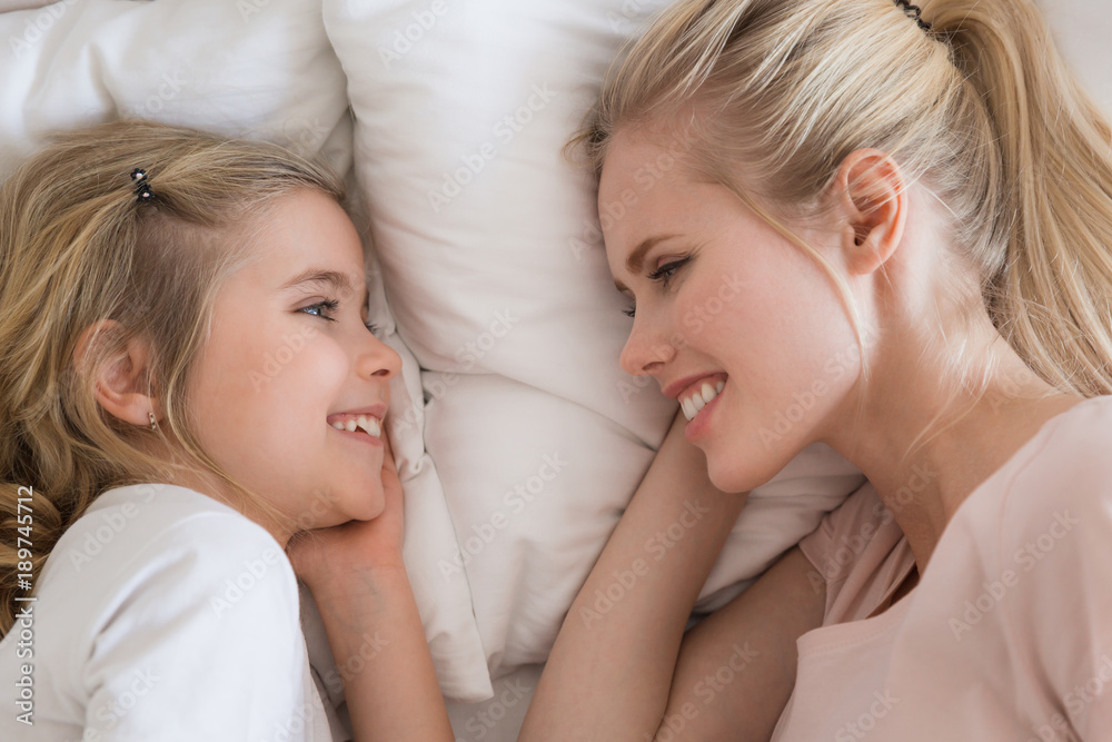 top view of smiling daughter and mother lying on bed and looking at each other