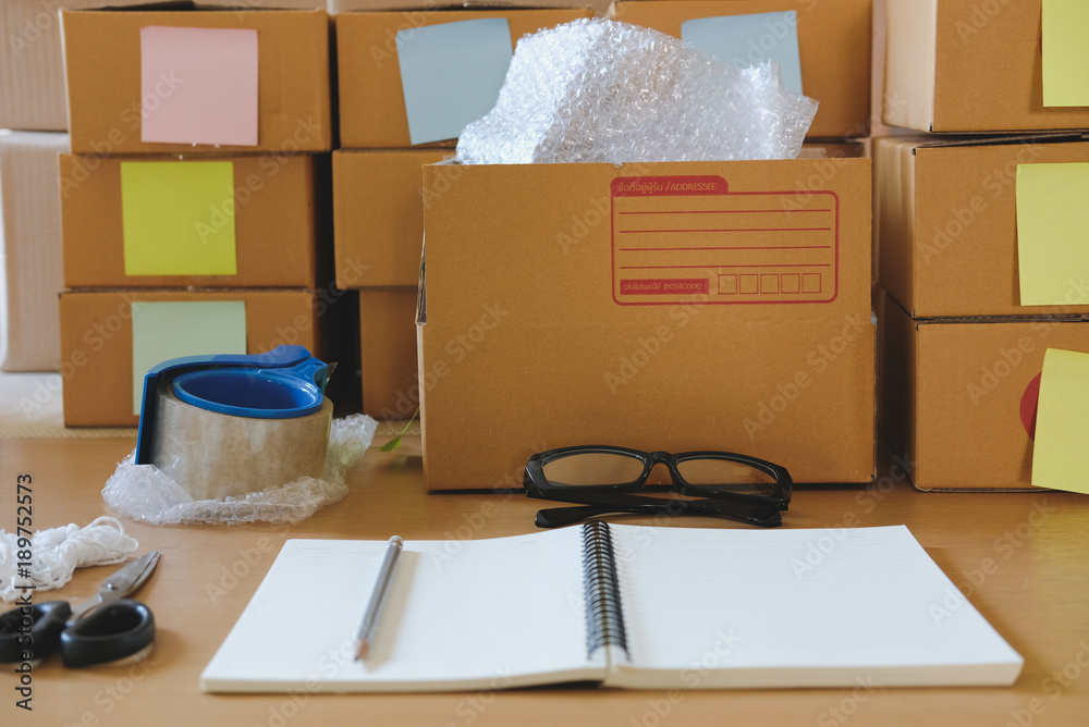 Fototapeta premium notebook, packing accessories at workplace of startup small business owner. cardboard parcel box for online selling. entrepreneur seller desk. e-commerce, shipping concept