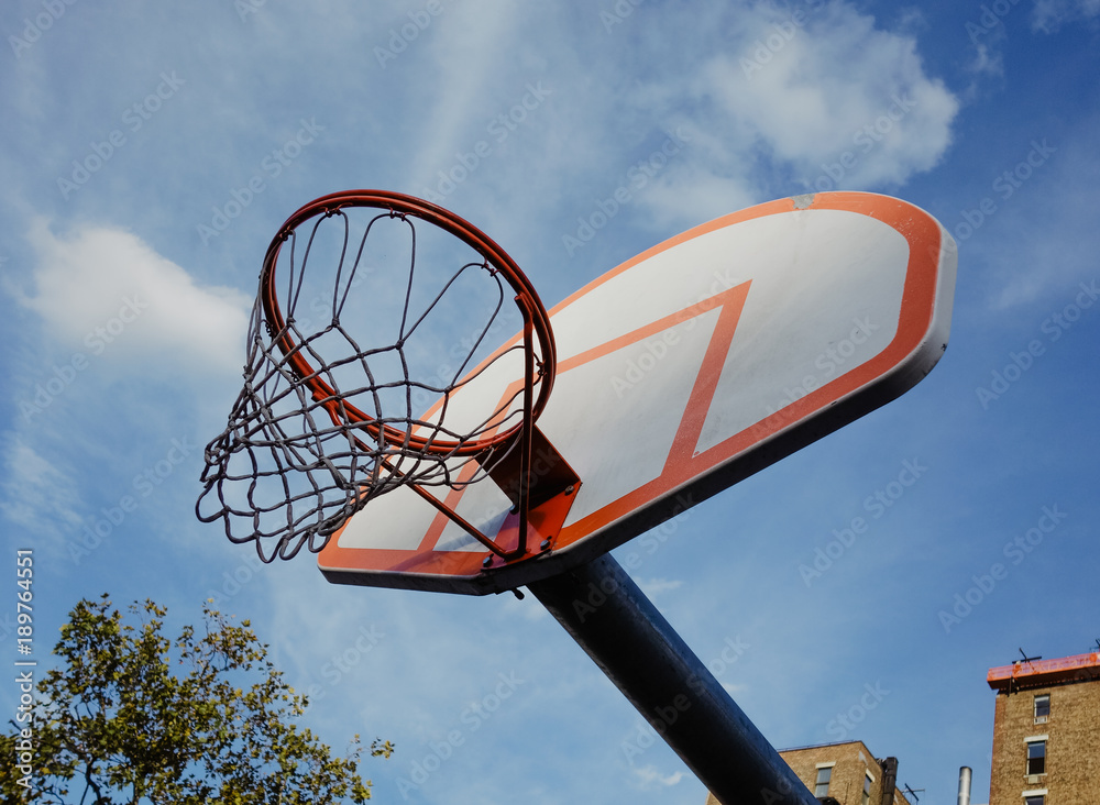 Basketball backboard with hoop and net in bright sunlight against blue sky with clouds and brickhouses in background in the Bronx neighbourhood in New York City, USA.