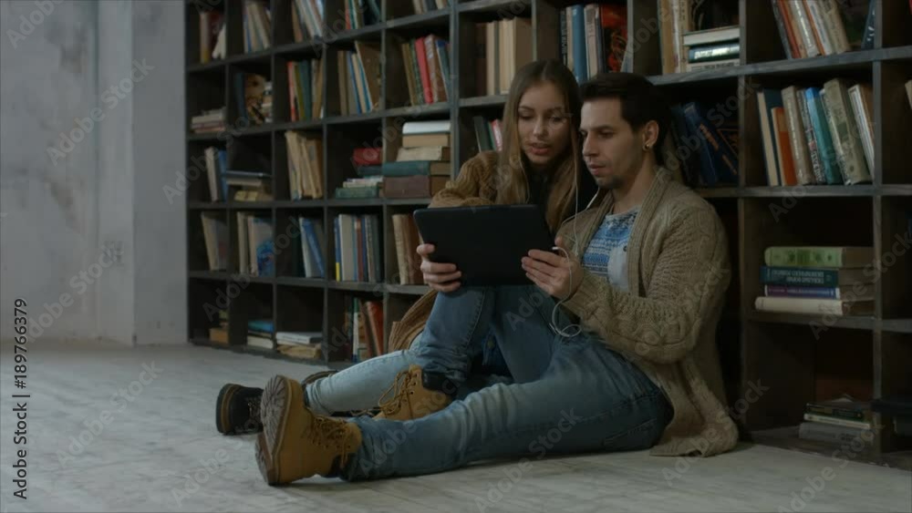 Smiling attractive couple in headphones working on digital tablet pc while sitting on floor in campus library over bookshelf background. Happy students with tablet pc studying in university library Smiling attractive couple in headphones working on digital tablet pc while sitting on floor in campus library over bookshelf background. Happy students with tablet pc studying in university library