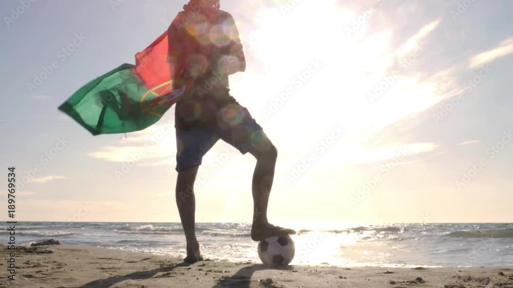 Young man wearing portuguese portugal national flag as super hero cape stands with foot on football on the sea shore sand looking at the ocean at the beach at sunset camera steadycam gimbal revolving