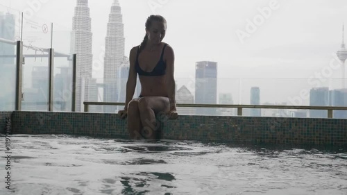 Pretty slim rich blonde woman in black bikini relaxing at the edge of roof top swimming pool or jakuzzi at resort or in hotel with view over city skyline. Success, vacation leisure activity concept. photo