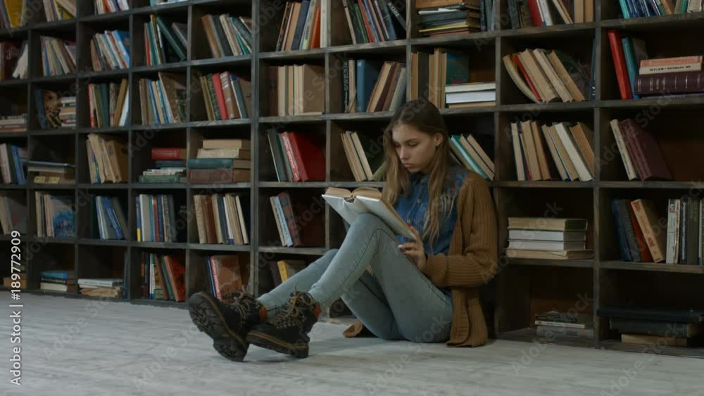 Frustrated female student has a lot to read in library. Exhausted female student covering her face with a book while sitting on the floor in college library and studying. Frustrated female student has a lot to read in library. Exhausted female student covering her face with a book while sitting on the floor in college library and studying.